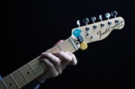 A close-up of a hand playing a Fender Telecaster guitar. The fingers are positioned on the fretboard, and there is a blue and yellow guitar pick holder attached to the headstock. The guitar is set against a dark background.