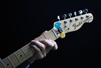 A close-up of a hand playing a Fender Telecaster guitar. The fingers are positioned on the fretboard, and there is a blue and yellow guitar pick holder attached to the headstock. The guitar is set against a dark background.