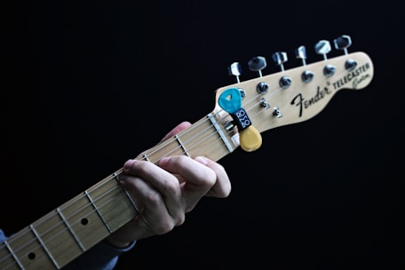A close-up of a hand playing a Fender Telecaster guitar. The fingers are positioned on the fretboard, and there is a blue and yellow guitar pick holder attached to the headstock. The guitar is set against a dark background.