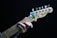 A close-up of a hand playing a Fender Telecaster guitar. The fingers are positioned on the fretboard, and there is a blue and yellow guitar pick holder attached to the headstock. The guitar is set against a dark background.