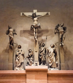A detailed sculpture depiction of the crucifixion of Jesus Christ, with Jesus shown on the cross flanked by two other figures also on crosses. Below are statues of lamenting figures, including Mary and several apostles, showcasing expressions of sorrow and reverence. The scene is dramatic and solemn with intricate details in the attire and facial expressions.