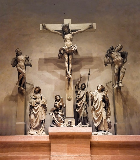A detailed sculpture depiction of the crucifixion of Jesus Christ, with Jesus shown on the cross flanked by two other figures also on crosses. Below are statues of lamenting figures, including Mary and several apostles, showcasing expressions of sorrow and reverence. The scene is dramatic and solemn with intricate details in the attire and facial expressions.