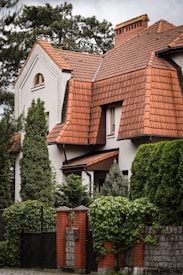 A residential building with unique architecture featuring steep, red-tiled roofs surrounded by dense greenery. The facade is primarily white with red brick accents, including a brick fence with climbing vines.