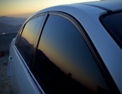 A close-up view of a car with dark tinted windows reflecting a mountainous landscape during sunset. The sky exhibits a gradient of warm colors transitioning into the silhouette of the mountain range in the background.