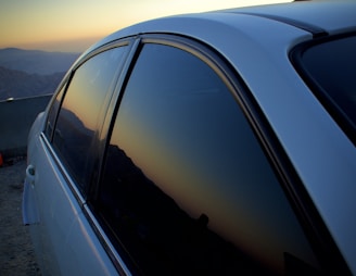 A close-up view of a car with dark tinted windows reflecting a mountainous landscape during sunset. The sky exhibits a gradient of warm colors transitioning into the silhouette of the mountain range in the background.
