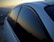 A close-up view of a car with dark tinted windows reflecting a mountainous landscape during sunset. The sky exhibits a gradient of warm colors transitioning into the silhouette of the mountain range in the background.