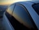 A close-up view of a car with dark tinted windows reflecting a mountainous landscape during sunset. The sky exhibits a gradient of warm colors transitioning into the silhouette of the mountain range in the background.