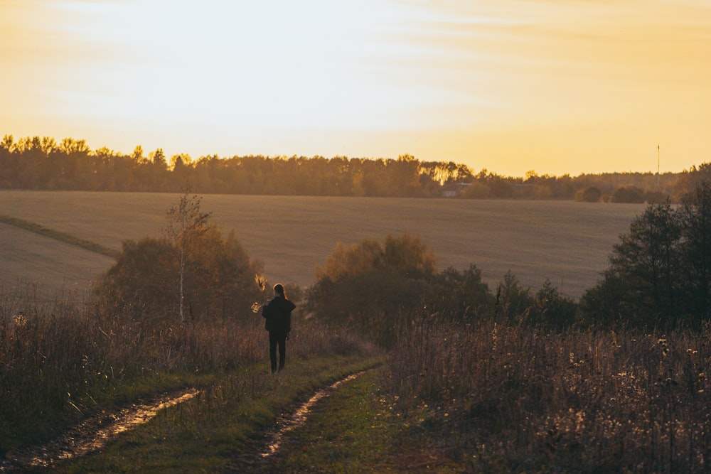 a person standing on a dirt path in a field with trees and a sunset