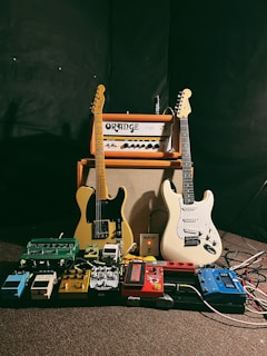 Two electric guitars stand upright in front of an Orange amplifier, surrounded by a variety of colorful guitar effect pedals on the floor. The background is dark, accentuating the vibrant colors of the instruments and pedals.