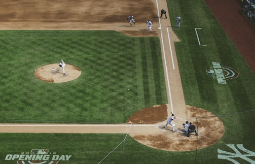 An aerial view of a baseball game in action, featuring a pitcher on the mound mid-throw to a batter at home plate. The catcher and umpire are positioned behind the batter, awaiting the pitch, while infielders are ready for the play. The field is lush and green, marked by dirt paths around the bases, and the field markings indicate 'Opening Day'.
