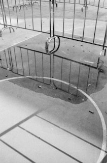 A black and white image depicting metal barriers on a paved surface. The ground is wet, creating reflections of the barriers in a puddle. The scene includes scattered leaves, and the lines of a sports court or playground are visible.