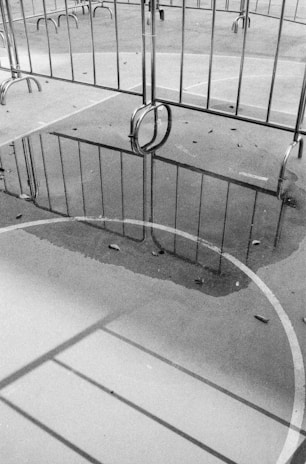 A black and white image depicting metal barriers on a paved surface. The ground is wet, creating reflections of the barriers in a puddle. The scene includes scattered leaves, and the lines of a sports court or playground are visible.