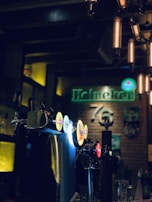 A dimly lit bar setting features several beer taps with illuminated logos. The background includes a sign with neon lighting and various bottles displayed on shelves. The ambiance is warm and inviting, with dark wood and metal accents reflecting a cozy atmosphere.