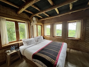 A cozy bedroom with rustic wooden elements, featuring a large bed covered with white linens and a decorative throw. The room is surrounded by windows offering lush green views outside, creating a natural and peaceful ambiance. A small wooden side table holds two bottles of water and some electrical devices, while wooden beams and woven walls enhance the natural aesthetic.
