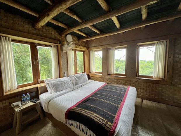 A cozy bedroom with rustic wooden elements, featuring a large bed covered with white linens and a decorative throw. The room is surrounded by windows offering lush green views outside, creating a natural and peaceful ambiance. A small wooden side table holds two bottles of water and some electrical devices, while wooden beams and woven walls enhance the natural aesthetic.
