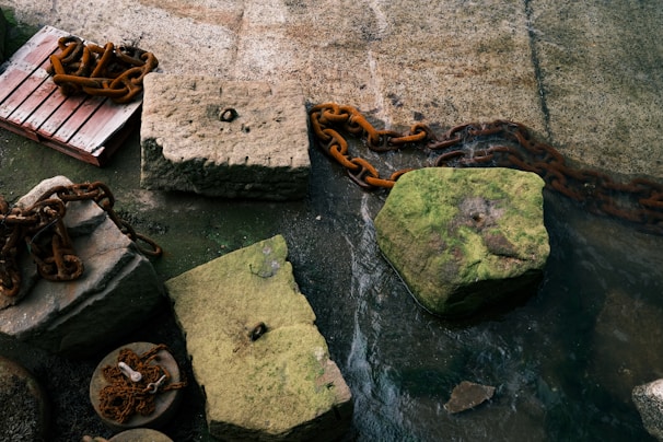 Large, weathered concrete blocks are connected by heavy, rusted chains. A red wooden pallet is partially visible, placed near one of the blocks. The surface surrounding the blocks is covered with moss and appears wet, suggesting proximity to water.
