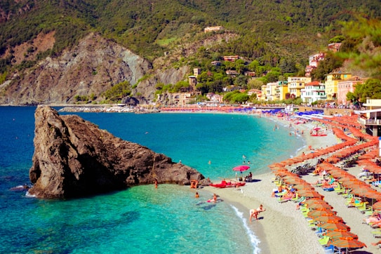 A picturesque beach scene with turquoise waters and a prominent rocky outcrop on the left. The sandy shore is lined with colorful beach umbrellas and sun loungers, with several people enjoying the sun and sea. In the background, there are charming buildings nestled against a lush, green hillside.