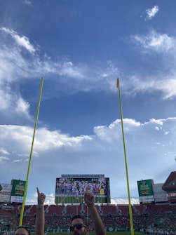 The image captures a view inside a large sports stadium, with bright yellow goalposts in the foreground reaching up towards a blue sky filled with fluffy white clouds. A crowd is present in the stands, partially visible underneath a large scoreboard displaying a sports game. Two spectators stand at the bottom center of the frame, with their hands raised, pointing upwards.