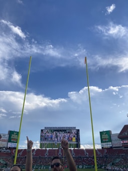 The image captures a view inside a large sports stadium, with bright yellow goalposts in the foreground reaching up towards a blue sky filled with fluffy white clouds. A crowd is present in the stands, partially visible underneath a large scoreboard displaying a sports game. Two spectators stand at the bottom center of the frame, with their hands raised, pointing upwards.