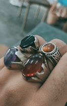 Four rings with large, colorful stones are being worn on a hand. The stones include a dark green, a purple, a deep red, and an amber-colored one. The metal settings appear to be silver.