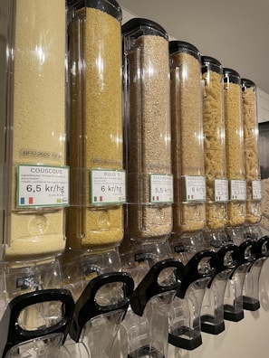 A row of transparent dispensers filled with various types of grains and pasta. Each dispenser is labeled with pricing information in kr/kg and the type of grain or pasta, such as couscous, bulgur, and macaroni. The dispensers are aligned and well-organized, equipped with black handles at the bottom for dispensing the contents.