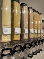 A row of transparent dispensers filled with various types of grains and pasta. Each dispenser is labeled with pricing information in kr/kg and the type of grain or pasta, such as couscous, bulgur, and macaroni. The dispensers are aligned and well-organized, equipped with black handles at the bottom for dispensing the contents.