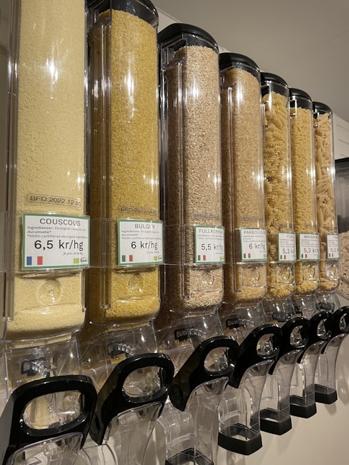 A row of transparent dispensers filled with various types of grains and pasta. Each dispenser is labeled with pricing information in kr/kg and the type of grain or pasta, such as couscous, bulgur, and macaroni. The dispensers are aligned and well-organized, equipped with black handles at the bottom for dispensing the contents.