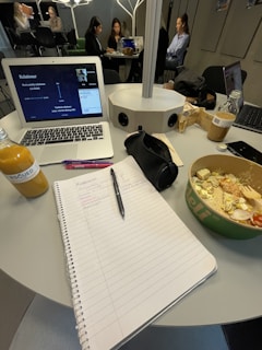 A workspace is depicted with a table holding a variety of items, including an open notebook with handwritten notes, a laptop showing a virtual meeting or presentation, a salad in a bowl, a bottle of juice, and various writing utensils. In the background, several people are seated around another table, possibly engaged in their own tasks or discussions.