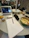 A workspace is depicted with a table holding a variety of items, including an open notebook with handwritten notes, a laptop showing a virtual meeting or presentation, a salad in a bowl, a bottle of juice, and various writing utensils. In the background, several people are seated around another table, possibly engaged in their own tasks or discussions.