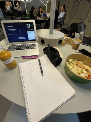 A workspace is depicted with a table holding a variety of items, including an open notebook with handwritten notes, a laptop showing a virtual meeting or presentation, a salad in a bowl, a bottle of juice, and various writing utensils. In the background, several people are seated around another table, possibly engaged in their own tasks or discussions.