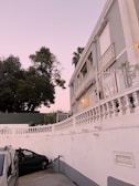 A residential building with ornate balconies and white railings stands next to a parking area where a black car and a silver vehicle are parked. The evening sky has a soft pink hue, and there are large green trees in the background, offering a serene atmosphere. A warm light illuminates the exterior of the building, highlighting its architectural details.