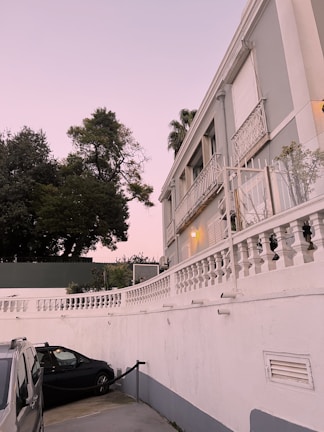 A residential building with ornate balconies and white railings stands next to a parking area where a black car and a silver vehicle are parked. The evening sky has a soft pink hue, and there are large green trees in the background, offering a serene atmosphere. A warm light illuminates the exterior of the building, highlighting its architectural details.