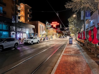 A nighttime street scene featuring a sign for 'Hotel Congress' with a temperature display. There are modern buildings on both sides of the street, illuminated by various lights. On the sidewalk to the right, there are red patio umbrellas, street lamps, and trees with festive lights. A few parked cars are visible on the left side of the road, and the street has tram tracks.