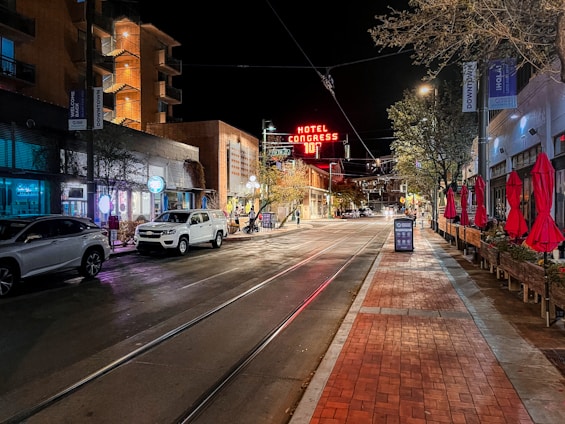 A nighttime street scene featuring a sign for 'Hotel Congress' with a temperature display. There are modern buildings on both sides of the street, illuminated by various lights. On the sidewalk to the right, there are red patio umbrellas, street lamps, and trees with festive lights. A few parked cars are visible on the left side of the road, and the street has tram tracks.