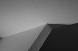 A minimalist black and white photograph featuring angular geometric shapes formed by intersecting lines and shadows. The image draws attention to the play of light and shadow, creating a stark contrast between light and dark areas.