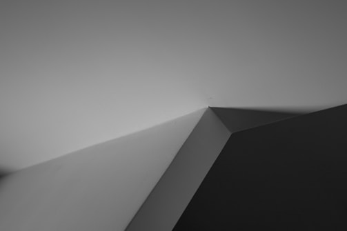 A minimalist black and white photograph featuring angular geometric shapes formed by intersecting lines and shadows. The image draws attention to the play of light and shadow, creating a stark contrast between light and dark areas.