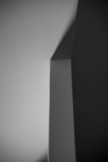 A minimalist black and white photograph featuring sharp geometric shapes and shadows in an abstract form. The image creates a strong contrast between light and dark areas, highlighting the clean lines and angles.