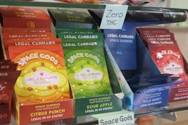 Packages of 'Space Gods' legal cannabis gummies are displayed on a shelf. The packaging includes vibrant colors like red, green, and blue. Labels detail various flavors, such as Citrus Punch, Sour Apple, and Wild Berry. A small sign reads 'Zero THC', indicating these are THC-free products.