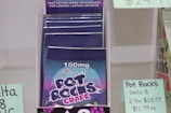 A display box contains packages labeled '100mg Pot Rocks Grape' with branding from Hemp Living. The color scheme involves dark purple and blue with a graphic of grapes and a hemp leaf icon. The signage indicates pricing for Delta 8 Pot Rocks at $12.99 each or two for $23.99.