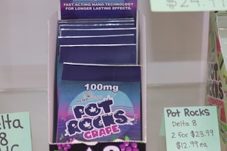 A display box contains packages labeled '100mg Pot Rocks Grape' with branding from Hemp Living. The color scheme involves dark purple and blue with a graphic of grapes and a hemp leaf icon. The signage indicates pricing for Delta 8 Pot Rocks at $12.99 each or two for $23.99.