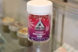 A container labeled 'Resin Series' featuring pink and purple branding with cannabis leaf designs is displayed on a glass surface. The product is named 'Pinkberry' and contains live resin, with details like 250mg potency and THC content mentioned on the label.