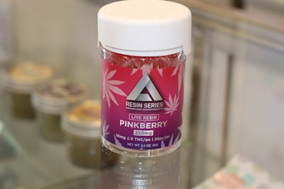 A container labeled 'Resin Series' featuring pink and purple branding with cannabis leaf designs is displayed on a glass surface. The product is named 'Pinkberry' and contains live resin, with details like 250mg potency and THC content mentioned on the label.