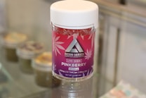 A container labeled 'Resin Series' featuring pink and purple branding with cannabis leaf designs is displayed on a glass surface. The product is named 'Pinkberry' and contains live resin, with details like 250mg potency and THC content mentioned on the label.