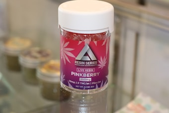 A container labeled 'Resin Series' featuring pink and purple branding with cannabis leaf designs is displayed on a glass surface. The product is named 'Pinkberry' and contains live resin, with details like 250mg potency and THC content mentioned on the label.