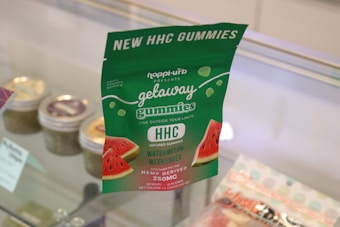A package of HHC gummies labeled 'getaway gummies' is prominently displayed. The packaging is green with white text and features images of watermelon slices. It advertises 'Watermelon Weekender' flavor and contains 250mg of hemp-derived content. Behind the package, jars and another sealed bag are visible in a glass display case.
