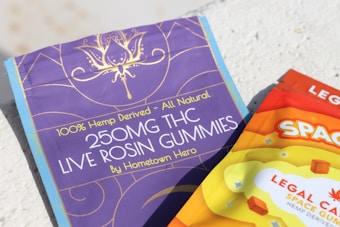Two packages of THC gummies are shown, one in purple and the other in orange. The purple package reads '250MG THC LIVE ROSIN GUMMIES - 100% Hemp-Derived - All Natural.' The orange package has bold letters spelling 'SPACE' with a colorful and vibrant design.