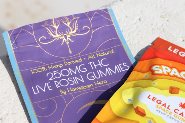 Two packages of THC gummies are shown, one in purple and the other in orange. The purple package reads '250MG THC LIVE ROSIN GUMMIES - 100% Hemp-Derived - All Natural.' The orange package has bold letters spelling 'SPACE' with a colorful and vibrant design.
