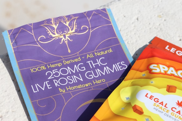 Two packages of THC gummies are shown, one in purple and the other in orange. The purple package reads '250MG THC LIVE ROSIN GUMMIES - 100% Hemp-Derived - All Natural.' The orange package has bold letters spelling 'SPACE' with a colorful and vibrant design.