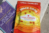 A brightly colored package of legal cannabis gummies labeled 'Space Gods' with a citrus punch flavor. The package displays information about its THC and CBD content, emphasizing that it's hemp-derived. The background shows another package partially visible, labeled as THC live rosin gummies.