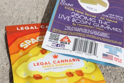 A close-up view of two packages of edible gummies on a textured surface. One package has a blue and purple design with information about 'Live Rosin Gummies,' including a 250mg THC content and '100% hemp derived' labeling. The other package is orange and yellow, labeled as 'Legal Cannabis Space Gummies' with 300mg THC content. Both packages have QR codes, branding, and legal information.
